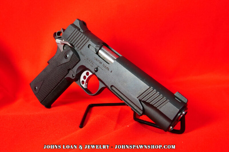 Matte black Kimber Custom TLE/RL II pistol in .45 ACP, shown on a display stand against a red backdrop, featuring diamond checkered grips, from John's Loan & Jewelry.