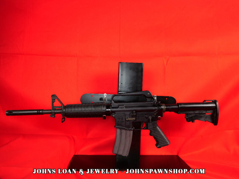 Olympic Arms CAR-15 semi-auto rifle, a vintage-style firearm, displayed against a red backdrop at John's Loan & Jewelry. The black AR-15 variant features a pinned flash hider and a 30+1 capacity magazine.
